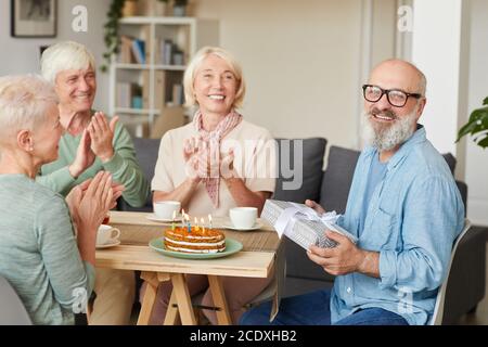 Portrait of senior bearded man holding gift box and smiling at camera while his friends congratulating him at the table Stock Photo