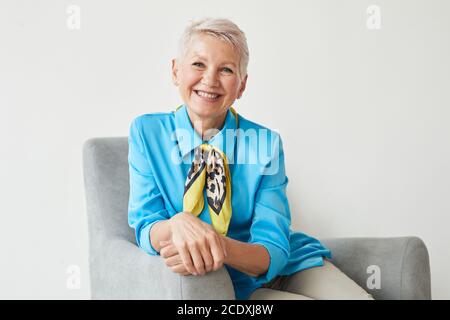 Portrait of elegant mature woman in beautiful blue blouse sitting on armchair and smiling at camera isolated on white background Stock Photo