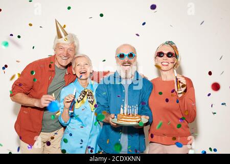 Portrait of group of senior people with birthday cake standing under the confetti and smiling at camera against the white background Stock Photo