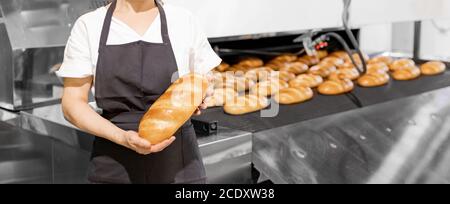 Close-up baker in apron holding hot baguette bread in hands on background of automated production line Stock Photo