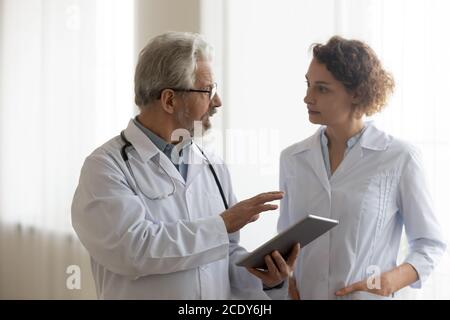 Serious mature doctor and young nurse talking, working together Stock Photo