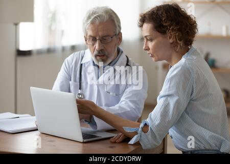 Serious mature doctor showing checkup result on laptop to patient Stock Photo