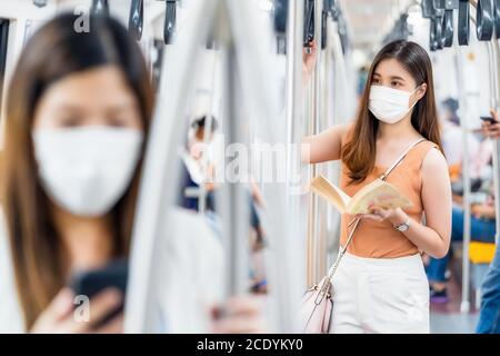 Young Asian woman passenger wearing surgical mask and reading the book in subway train when traveling in big city at Covid19 outbreak, Infection and P Stock Photo