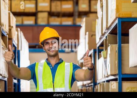 Portrait of Indian warehouse worker man with safety clothes standing with confident and showing thumps up in local workplace with warehouse or factory Stock Photo