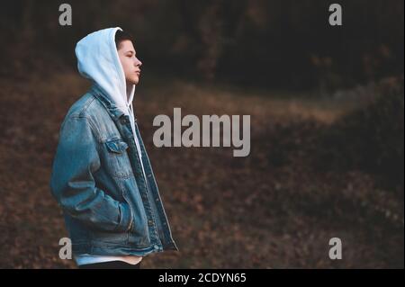 Handsome teen boy 15-16 year old wearing denim jacket and white hoodie standing outdoors over nature autumn background. Teenagerhood. Stock Photo