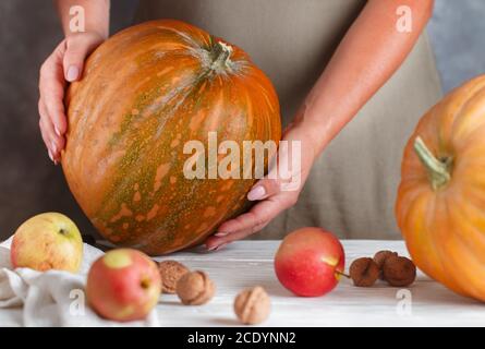 Whole large organic pumpkin in women's hands, ripe apples and walnuts on the table. Thanksgiving, Halloween. Autumn harvest. Selective focus Stock Photo