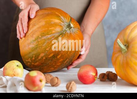 Whole large organic pumpkin in women's hands, ripe apples and walnuts on the table. Thanksgiving, Halloween. Autumn harvest. Selective focus Stock Photo