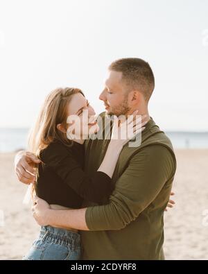 Young adult couple standing on beach, looking each other. Handsome man embracing beautiful laughing woman. Millennial male and female posing on coast on sunny day and looking away Stock Photo