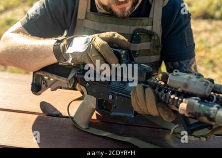 Close-up of bearded man in gloves sitting at table outdoors and assembling rifle Stock Photo