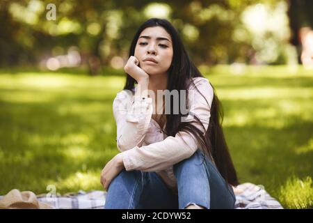 Summertime Sadness. Portrait Of Thoughtful Asian Woman Sitting On Plaid In Park Stock Photo