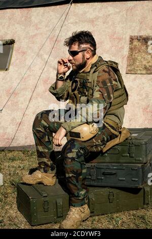 Pensive bearded man in camouflage outfit sitting on metal suitcases and smoking cigarette outdoors Stock Photo