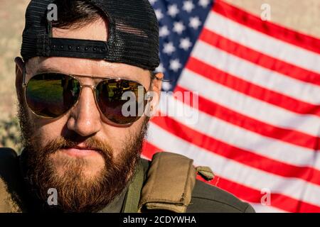 Portrait of serious brutal American man with beard wearing sunglasses and cap standing against national flag Stock Photo