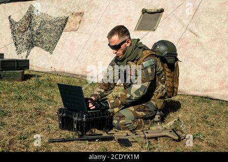 Concentrated young programmer soldier sitting on ground and working with laptop while analyzing army route Stock Photo