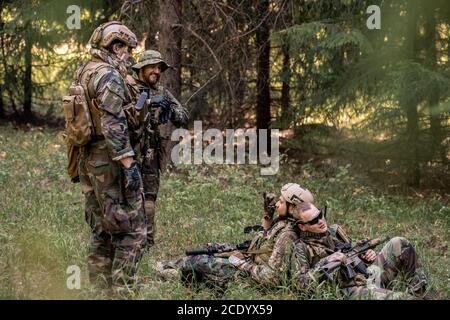 Young soldiers sitting back to back on grass and talking to colleagues while resting in forest Stock Photo