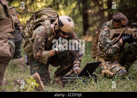 Busy soldier with backpack using military laptop while passing information through radio device in forest Stock Photo