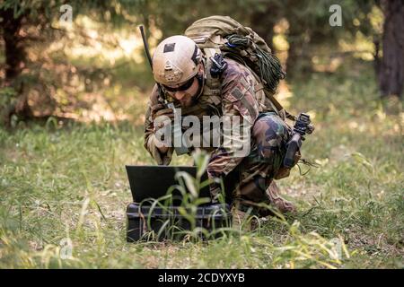 Bearded soldier in camouflage uniform sitting in forest and sending message through radio device while controlling military operation using computer Stock Photo