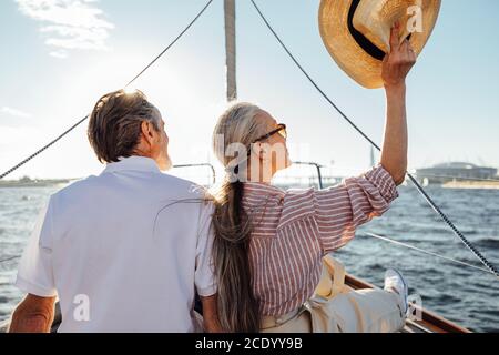 Back view of relaxed mature couple sitting on yacht. Stylish woman waving hat during boat trip. Stock Photo
