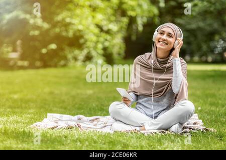 Happy woman in hijab enjoying music while resting at park Stock Photo