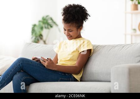 African Teen Girl Using Phone Sitting On Sofa At Home Stock Photo