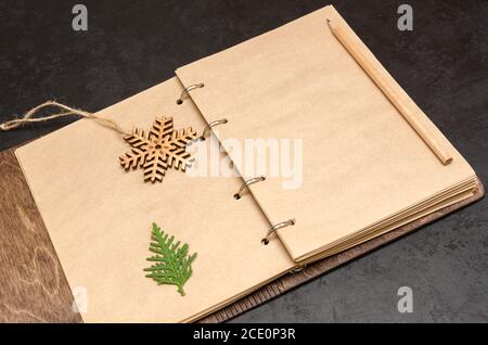 Notebook in wooden cover with kraft blank sheets, pencil, fir tree branch and wooden snowflake on black background. Plan next year concept. To do list Stock Photo