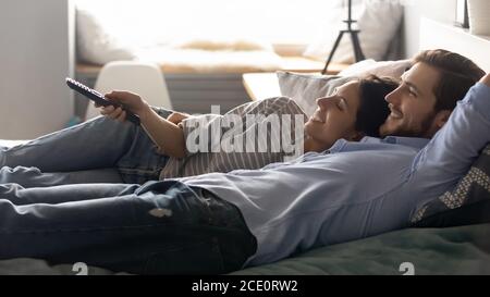 Happy relaxed couple lying on bed, using remote tv controller. Stock Photo