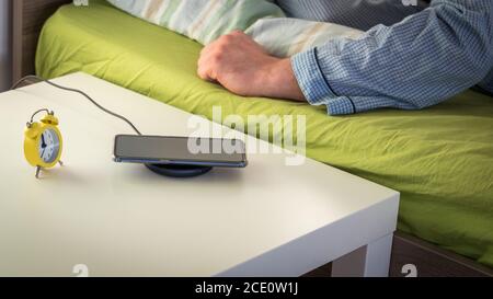 A charging phone on the bedside table next to the bed and a sleeping man Stock Photo