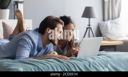 Interested young couple lying on bed, looking at computer screen. Stock Photo