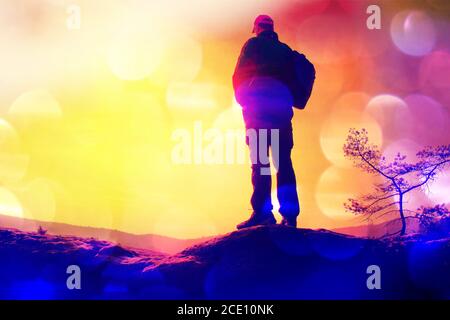 Film grain. Silhouette of man with red cap and  poles in hand. Man with big backpack stand on rock. Sunny spring daybreak in roc Stock Photo