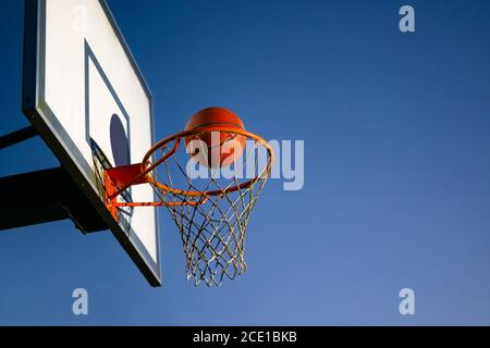 Street basketball ball falling into the hoop. Close up of orange ball above the hoop net with blue sky in the background. Concept of success, scoring Stock Photo