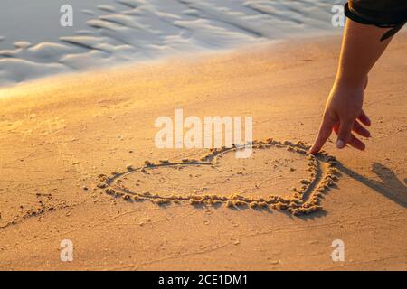 Close up view of woman hand drawing heart with finger on the sand at the beach. Romantic love symbol at tropical seashore at sunset Stock Photo