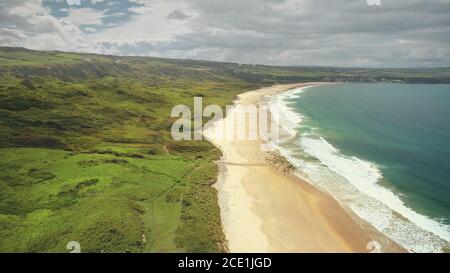 Aerial coast beach: Atlantic Ocean, Antrim county, Northern Ireland. People walking on sandy white shore with tranquil coastal wavy water. Cloudy summer scenery shot Stock Photo