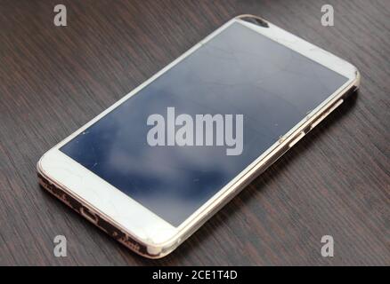 White smartphone with broken screen on wooden table. Dropped and old phone with reflection of the sky. Stock Photo