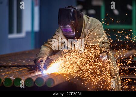 Welder in work in factory. Welding steel pipes on manufacturing (producing) plant. Sparks and smoke on dark background. Stock Photo