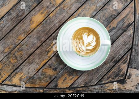 Cup of fresh creamy cappuccino with latte art on foam. Background of wooden table with shabby aged surface. Directly from above. Stock Photo