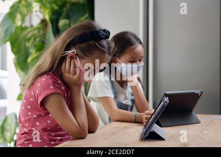 Multi ethnic schoolgirls wear facemasks sit at desk look at tablet screen feels troubled think over task study remotely. Alpha generation gadget overu Stock Photo