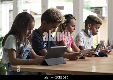 Four diverse kids wear facemasks sit at table use wireless gadgets ignoring each other prefer internet games and virtual communication. Alpha generati Stock Photo