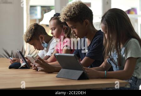 Four diverse kids wear facemasks sit at table use wireless gadgets ignoring each other prefer internet games and virtual communication. Alpha generati Stock Photo