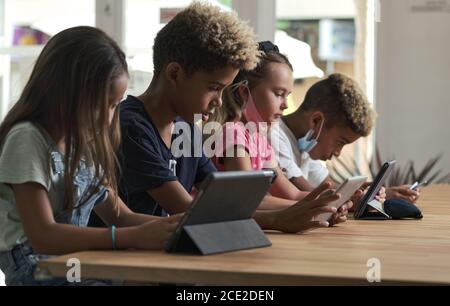 Four diverse kids wear facemasks sit at table use wireless gadgets ignoring each other prefer internet games and virtual communication. Alpha generati Stock Photo