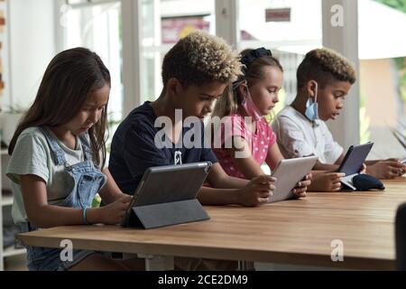 Four diverse kids wear facemasks sit at table use wireless gadgets ignoring each other prefer internet games and virtual communication. Alpha generati Stock Photo