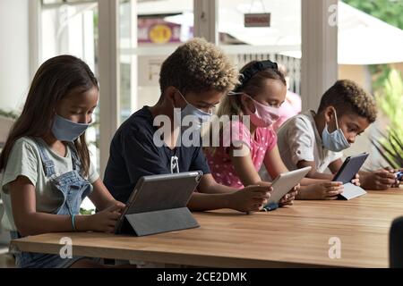 Four diverse kids wear facemasks sit at table use wireless gadgets ignoring each other prefer internet games and virtual communication. Alpha generati Stock Photo