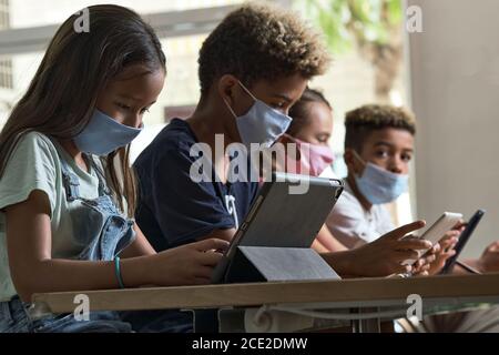 Four diverse kids wear facemasks sit at table use wireless gadgets ignoring each other prefer internet games and virtual communication. Alpha generati Stock Photo