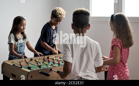 Group of multi cultural cute children stand indoor spend playtime with friends playing foosball together. Tabletop version of soccer lovers.