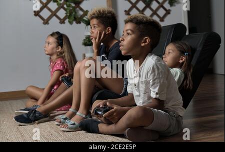 Four pretty multi national kids play to play station seated together at home. Alpha generation offspring addicted with virtual games problem concept Stock Photo