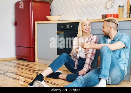 Romantic happy couple sitting on floor in kitchen and toasting champagne glasses Stock Photo