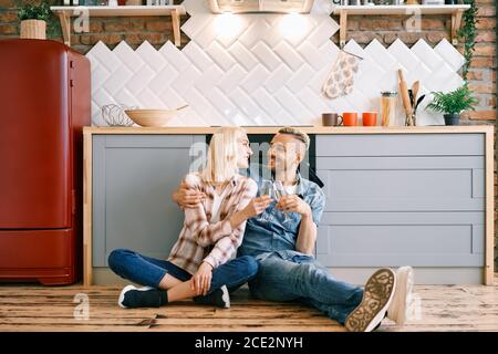 Romantic happy couple sitting on floor in kitchen and toasting champagne glasses Stock Photo