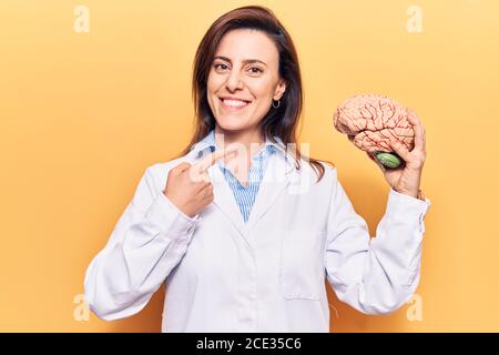 Young beautiful woman wearing doctor coat holding brain smiling happy pointing with hand and finger Stock Photo