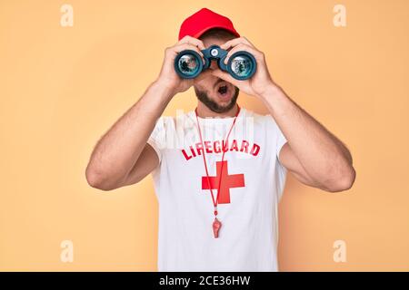 Young caucasian man wearing lifeguard t-shirt using binoculars in shock face, looking skeptical and sarcastic, surprised with open mouth Stock Photo