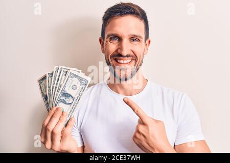 Young handsome man holding bunch of dollars banknotes over isolated white background smiling happy pointing with hand and finger Stock Photo