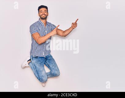 Young handsome hispanic man wearing casual clothes smiling happy. Jumping with smile on face pointing with fingers to the side over isolated white bac Stock Photo