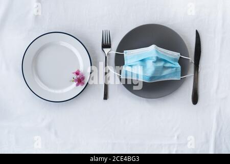 blue protection face mask for corona virus or covid 19 prevention on grey ceramic food dish with knife and fork and pink flower  for dinner in restaur Stock Photo